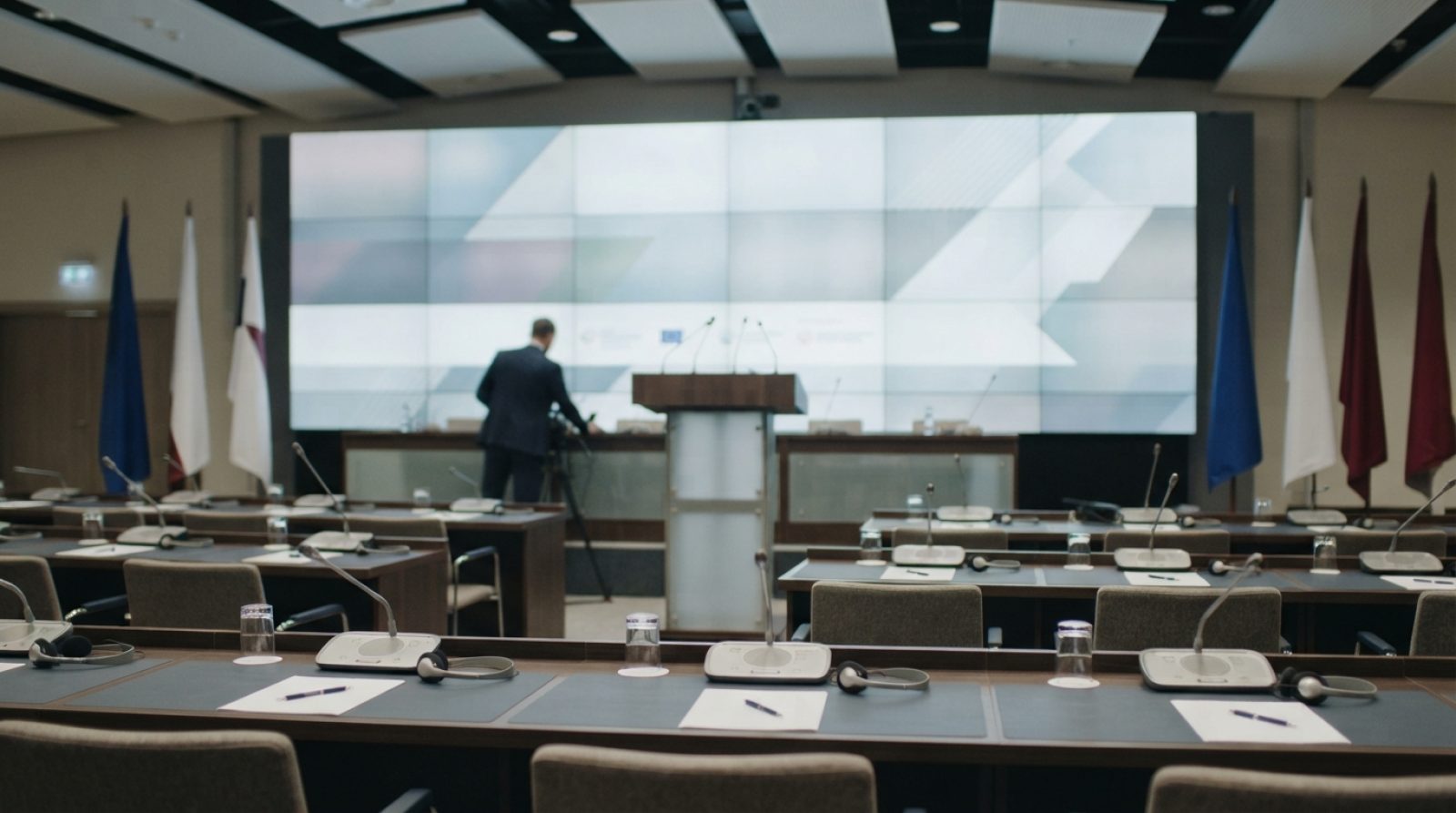 Illustration: A modern wide conference hall at a major international convention centre with an empty podium, orderly rows of desks equipped with trans