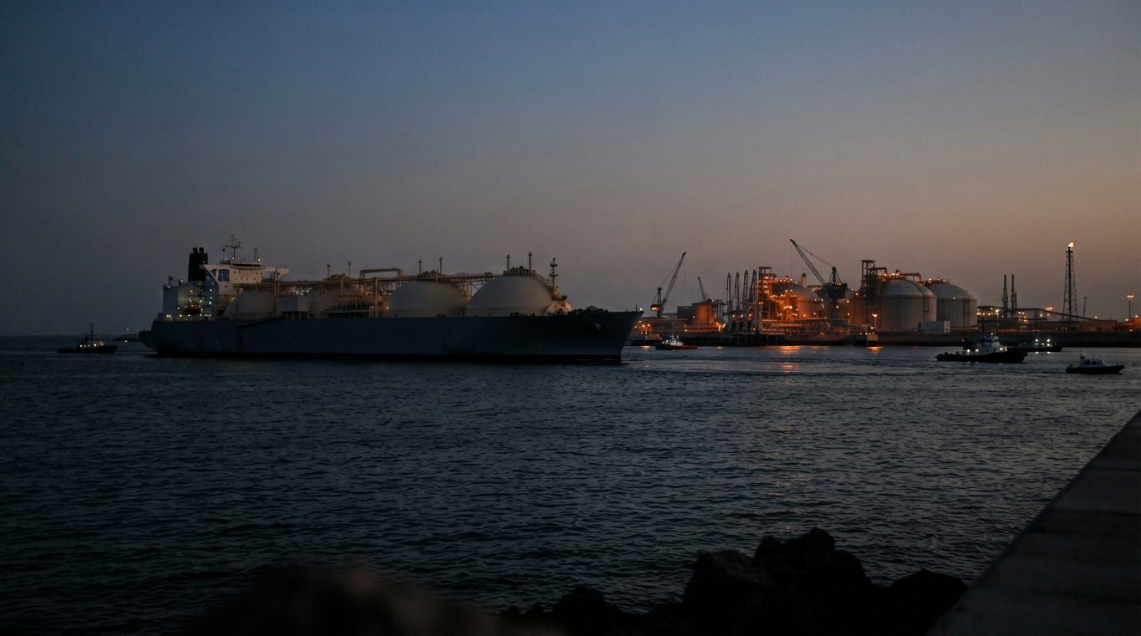 Illustration: A large commercial liquefied natural gas tanker vessel navigating coastal waters near an offshore energy loading terminal at twilight, w
