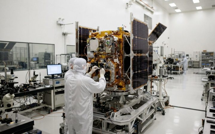 Illustration: Clean room satellite assembly laboratory with high-tech testing equipment, solar panel components, and optical instruments under bright