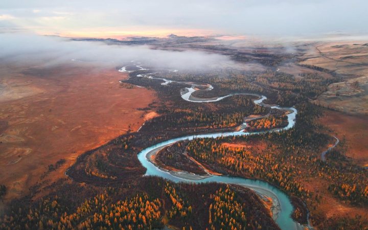 Aerial view of a winding turquoise river through an autumn forest landscape shrouded in morning mist