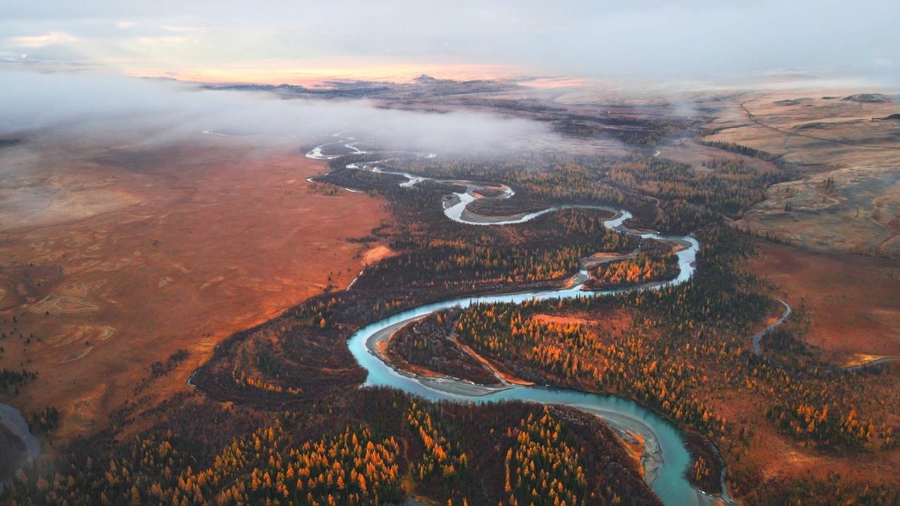 Aerial view of a winding river meandering through an autumn landscape beneath morning mist