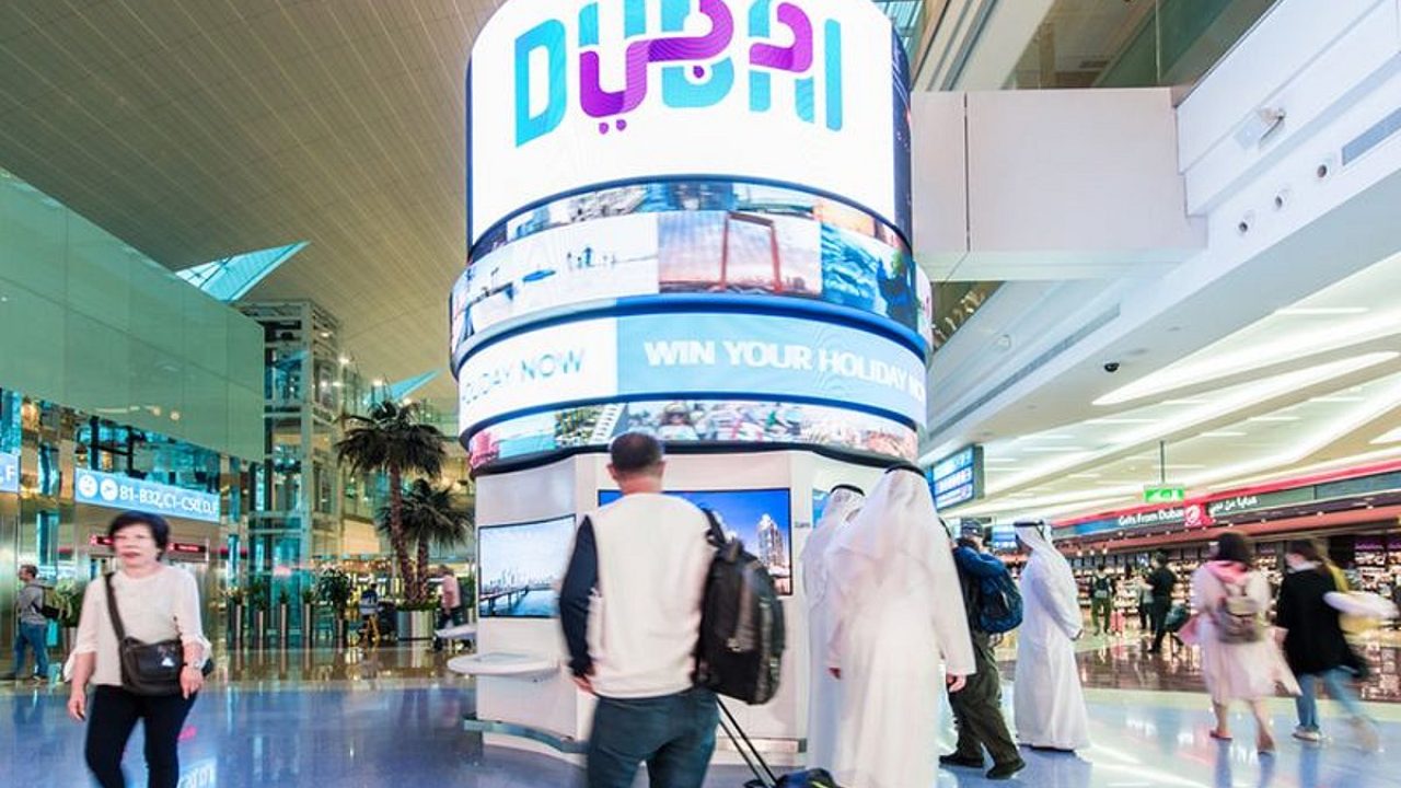Travelers walking past a large cylindrical digital Dubai display inside an airport terminal