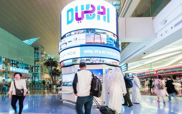 Travelers walking past a large cylindrical digital Dubai display inside an airport terminal