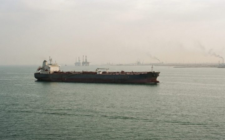 Illustration: A commercial oil tanker sailing on the Persian Gulf waters near the coastline under hazy daylight, with calm sea waves and distant indus
