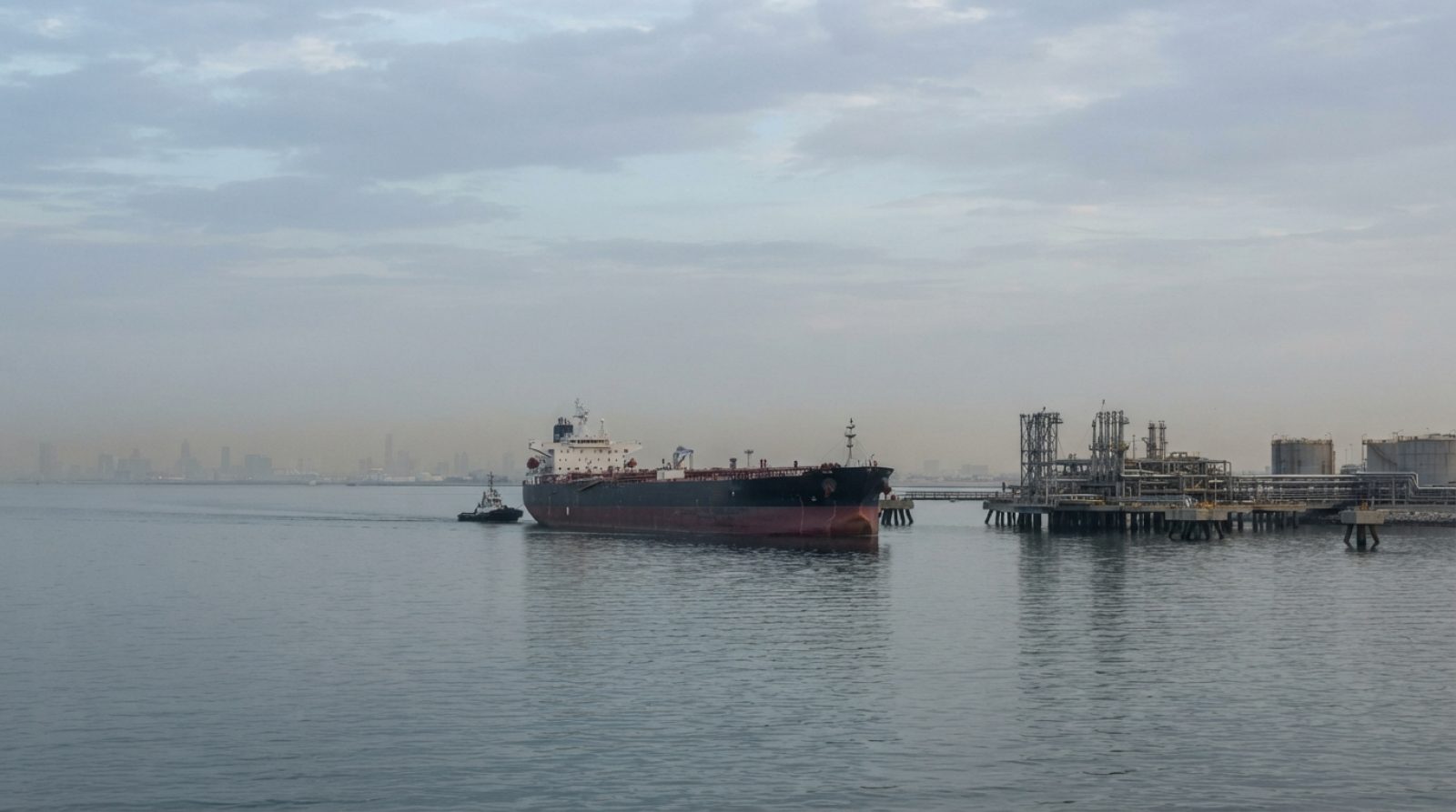 Illustration: An oil tanker navigating calm waters near an offshore terminal in the Persian Gulf under an overcast sky at dawn, with distant industria