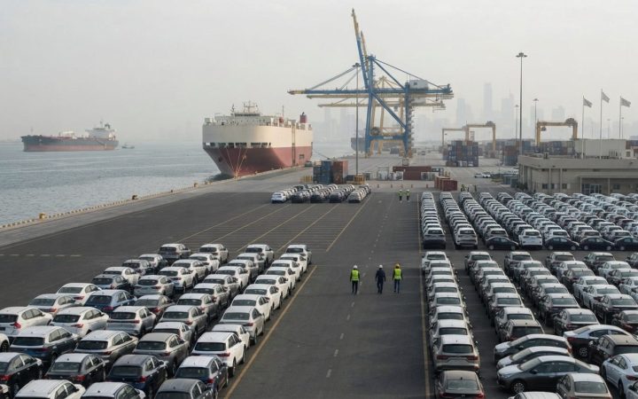Illustration: A commercial cargo terminal with rows of newly manufactured cars parked near a deepwater dock, with an oil tanker vessel visible on the