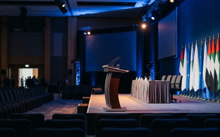 Illustration: An empty modern convention hall stage in Abu Dhabi with dramatic blue and amber stage lighting, professional conference seating, and pod