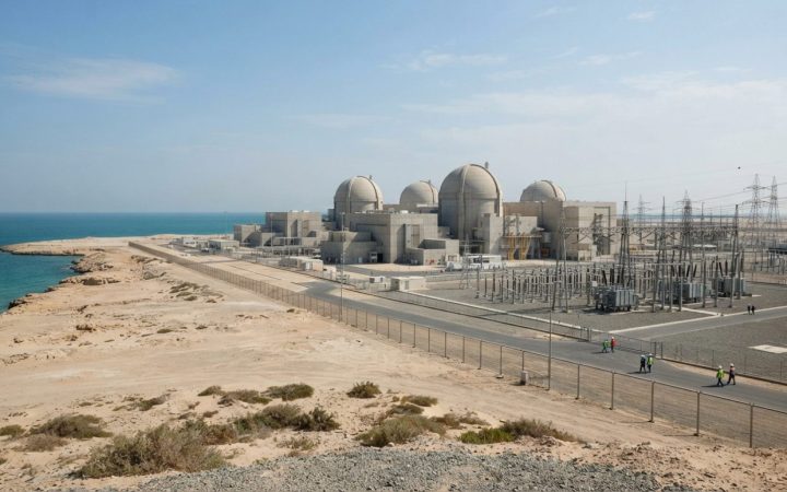 Illustration: A wide editorial view of a modern nuclear power plant facility with concrete containment domes and electrical distribution sub-stations