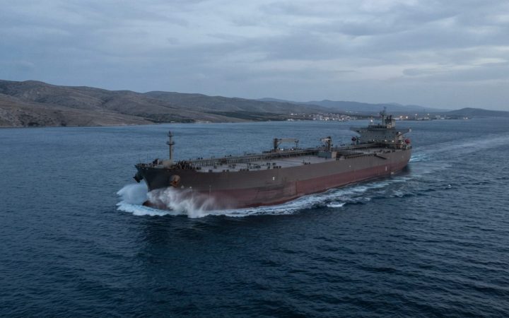 Illustration: A wide-angle realistic editorial photograph of a commercial crude oil supertanker sailing across deep blue water in a coastal strait at