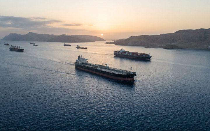 Illustration: An aerial editorial photograph of commercial supertankers and container vessels navigating calm deep blue waters near the Strait of Horm