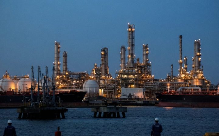 Illustration: A modern coastal petrochemical refinery complex at dusk with illuminated cracking towers, storage tanks, and industrial piping near a de
