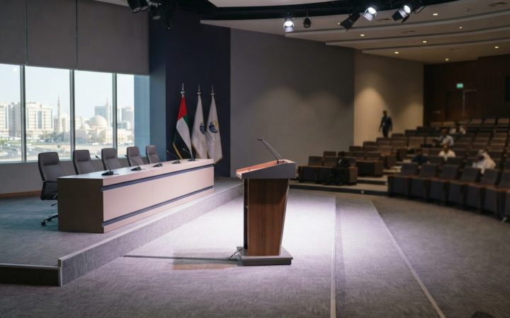 Illustration: A modern, bright business conference hall with empty stage seating, podium, and tiered audience rows illuminated by professional venue l
