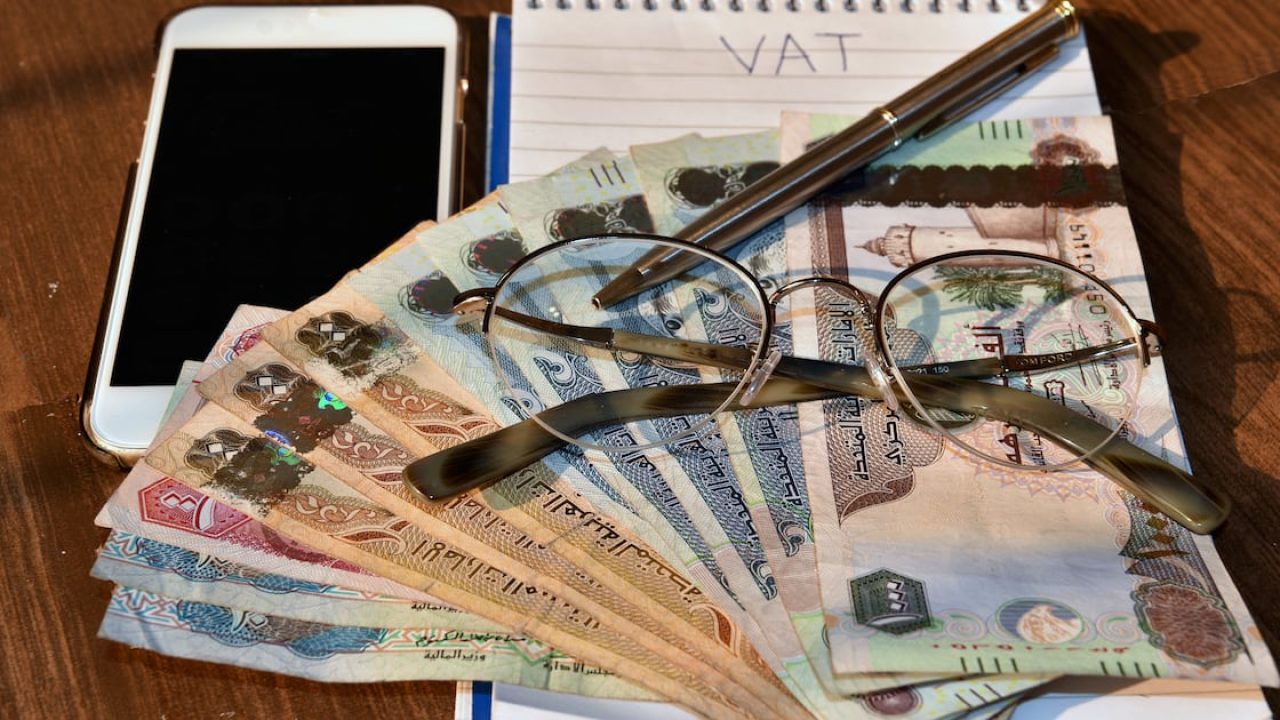 UAE dirham banknotes arranged with eyeglasses, a pen, a smartphone, and a notepad reading VAT