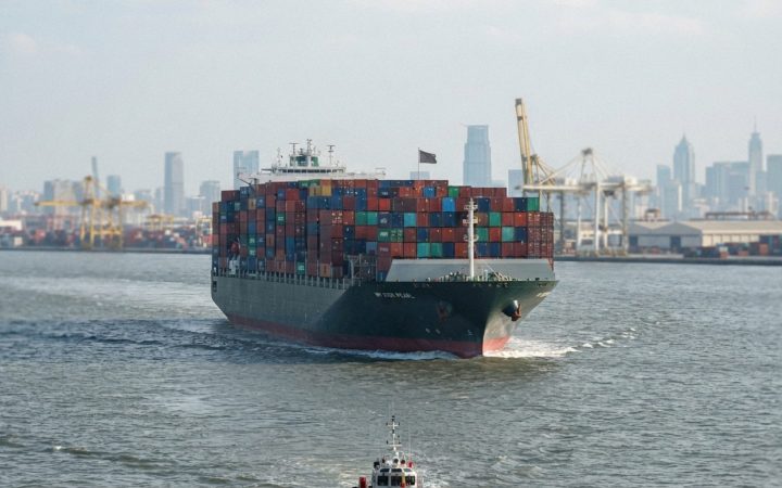 Illustration: A large commercial container ship loaded with colorful shipping containers moving through a wide maritime strait under daylight, viewed