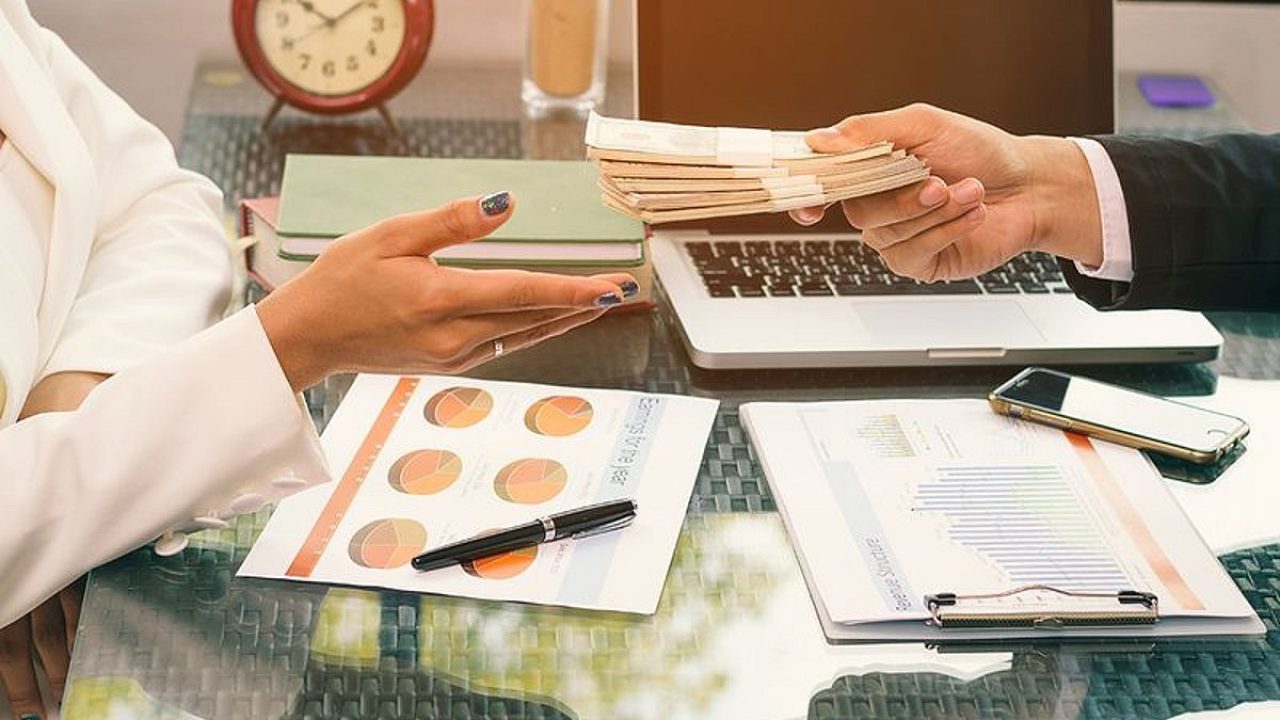 Businessperson handing a stack of banknotes across an office desk with charts and a laptop
