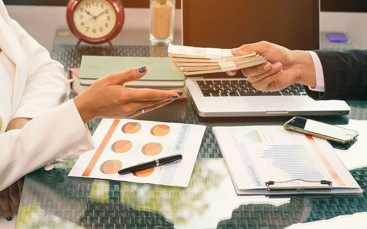 رجل أعمال يسلّم حزمة من الأوراق النقدية عبر مكتب عمل يضم رسوماً بيانية وحاسوباً محمولاً