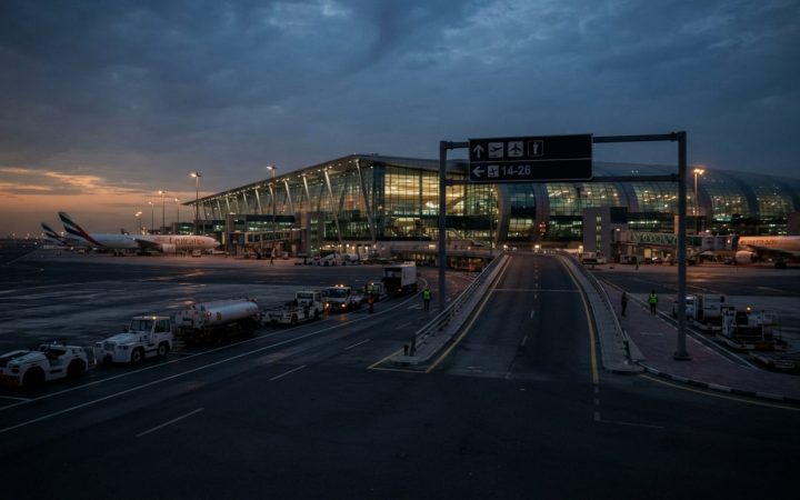 Illustration: Editorial photograph of a modern Middle Eastern international airport terminal exterior at dusk, showing empty tarmac access roads, dire